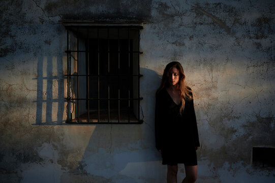 Full body of young female in casual black dress and boots leaning back against shabby stone wall of aged building with small window