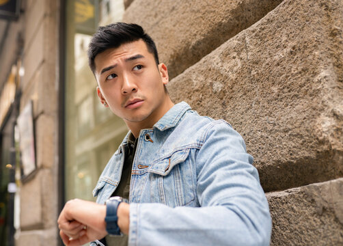 Low angle of serious Asian male in casual outfit and with wristwatch standing on street and looking away while waiting for appointment in city