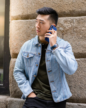 Serious Young Asian Male In Casual Outfit Speaking On Mobile Phone While Standing Near Stone Wall Of Urban Building