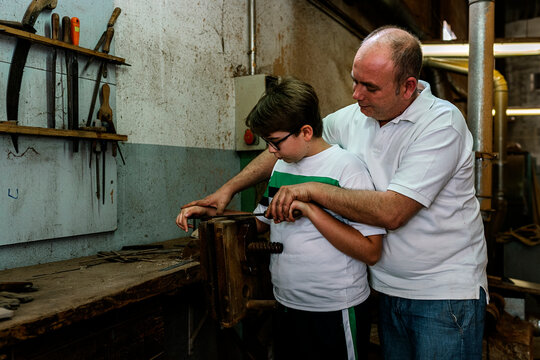 Side view of father teaching son using metal rasp for grinding wood in grungy dirty woodwork workshop