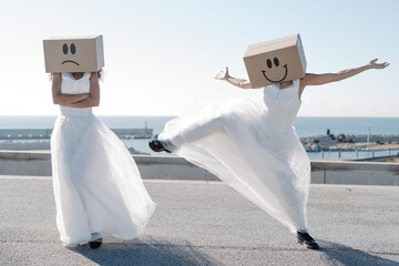 Unrecognizable women wearing white bridal dresses and carton boxes with happy and sad smileys on sunny rooftop