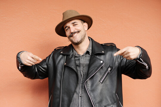 Trendy friendly male in hat and leather jacket smiling and looking at camera on beige background