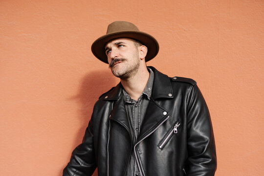 Trendy thoughtful male in hat and leather jacket on beige background looking away