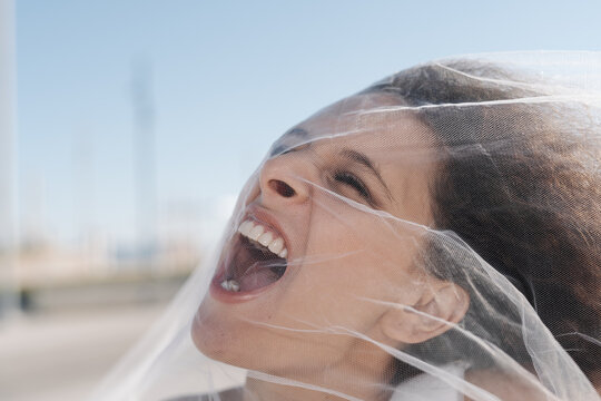 Young angry female with curly hair screaming through transparent veil on sunny clear day
