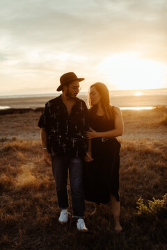 Full Body Young Couple In Casual Clothing Standing Holding Hands Through Meadow Covered With Yellow Dry Grass At Dawn Looking At Each Other