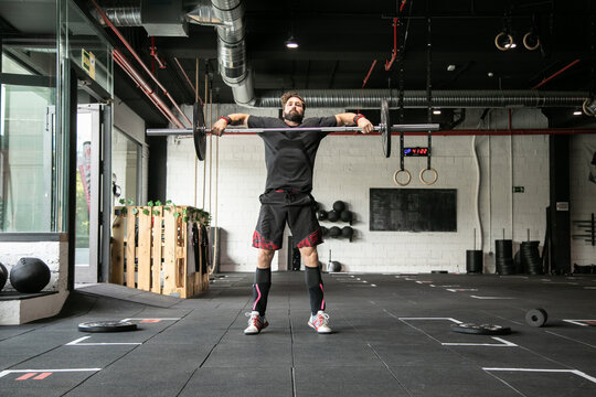 Muscular male athlete doing clean and jerk exercise with barbell during weightlifting training in gym looking at camera