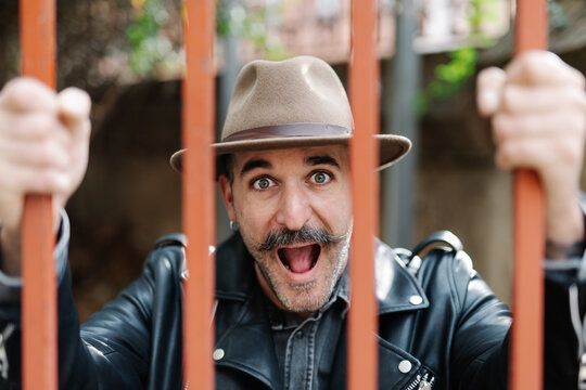 Trendy stylish male with mouth opened clinging metal fence being in capture and looking at camera