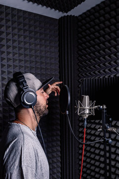 Side view of male singer in headphones and hat standing in acoustic room with soundproof walls and microphone and recording song in studio