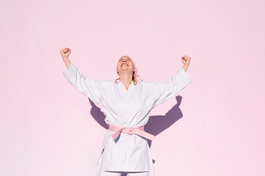 Determined mature woman in pink head cover and belt fighting karate in cancer battle concept doing victory gesture in the street on pink wall looking up