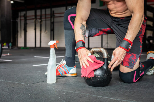 Unrecognizable strong male athlete wiping kettlebell with antibacterial liquid before training in gym during COVID 19 epidemic