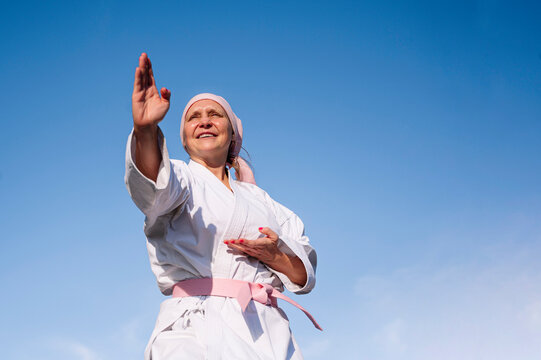 From below determined mature woman in pink head cover and belt fighting karate in cancer battle concept standing on blue sky background looking away