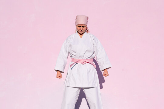 Determined thoughtful mature woman in pink head cover and belt fighting karate in cancer battle concept in the street on pink wall with eyes closed