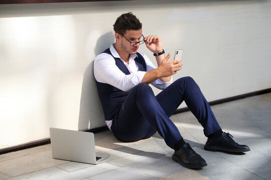 From above of young masculine office worker in formal suit and eyewear sitting on floor near wall with cellphone and netbook