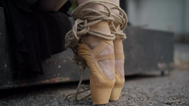 Camera moves down along slim ballerina legs bound with rope. Unrecognizable young Caucasian woman sitting outdoors. Concept of dancer tied to profession.