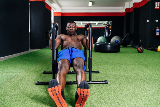 Muscular African American male with naked torso doing exercises on bars in gym while pumping biceps during intense training