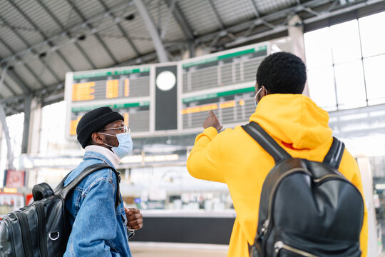 Traveling ethnic male friends with backpacks and in medical masks checking information on train schedule at station before departure during coronavirus epidemic