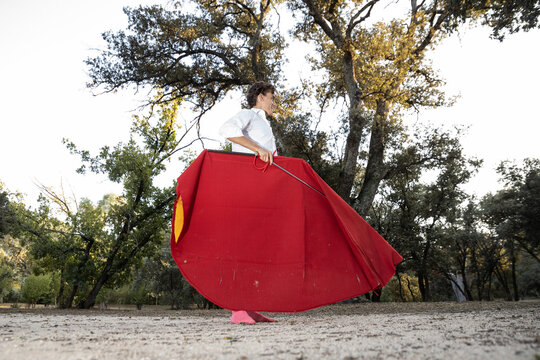From below side view of happy slim male bullfighter in red pants and white shirt holding cloak performing gracefully before corrida in nature