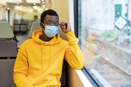 African American male tourist in mask sitting on passenger seat in contemporary train and looking out of window while traveling during coronavirus epidemic