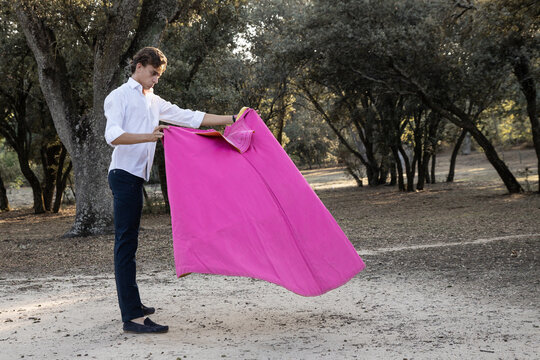 Side view of slim male bullfighter in red pants and white shirt holding cloak performing gracefully before corrida in nature