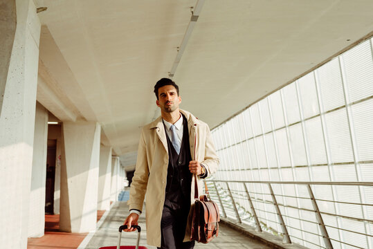Serious man in stylish clothes with suitcase walking along corridor at airport at daytime
