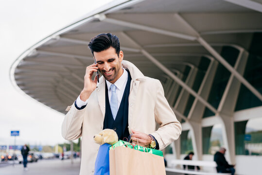 Positive man with black hair in formal clothes standing with shopping bags and talking on phone against modern building