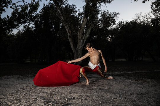 Side view of slender male bullfighter in red costume and with vivid cloak performing alone in nature in evening