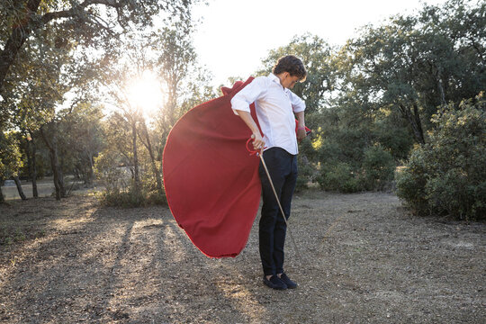 Slim focused male bullfighter in black pants and white shirt holding red cloak performing gracefully before corrida in nature
