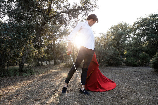 Slim unrecognizable male bullfighter in black pants and white shirt holding red cloak performing gracefully before corrida in nature