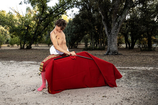 Focused slim male shirtless bullfighter in red pants holding cloak performing gracefully before corrida in nature