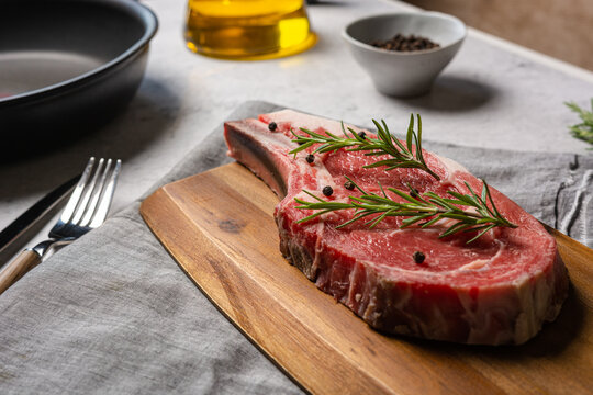 Uncooked t bone beef steak garnished with black pepper and rosemary sprigs placed on wooden chopping board in kitchen