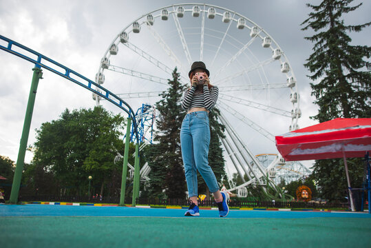 Low angle of young hipster female traveler in trendy outfit and hat covering face with instant camera while capturing pictures in amusement park against Ferris wheel during summer weekend in Saint Petersburg