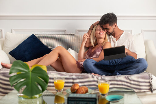 Happy young multiracial couple cuddling and browsing tablet while sitting together on cozy sofa near table with breakfast in morning - Powered by Adobe