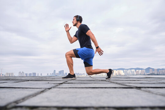 Ground level side view of athletic male in headphones listening to music and doing lunges during workout in city