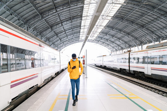 Ethnic male traveler with backpack and in mask standing on platform with train and using cellphone while waiting for departure during COVID 19 epidemic
