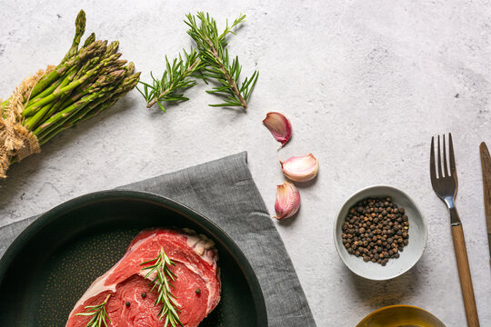 From above uncooked raw t bone steak garnished with rosemary and black pepper peas placed in frying pan on table in kitchen