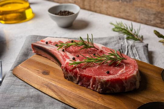 Uncooked t bone beef steak garnished with black pepper and rosemary sprigs placed on wooden chopping board in kitchen