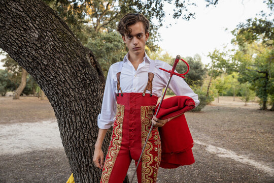 Young matador wearing red traditional costume standing in nature with cape and sword looking at camera