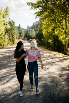 Cheerful young female friends in casual wear and protective masks hugging and having fun while standing on asphalt country road leading through forest in autumn day