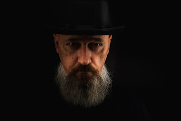 portrait of a young, handsome, emotional, serious man with a gray long beard and a black hat on his head, isolated on a dark background. There is a place for an inscription