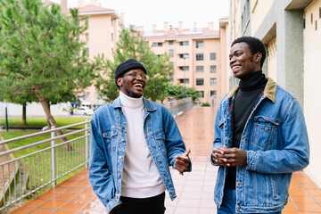 Cheerful African American male friends in stylish denim clothes standing on street and laughing while telling jokes and having fun