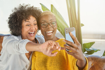 Cheerful African American females in love hugging in armchair and taking selfie on mobile phone camera