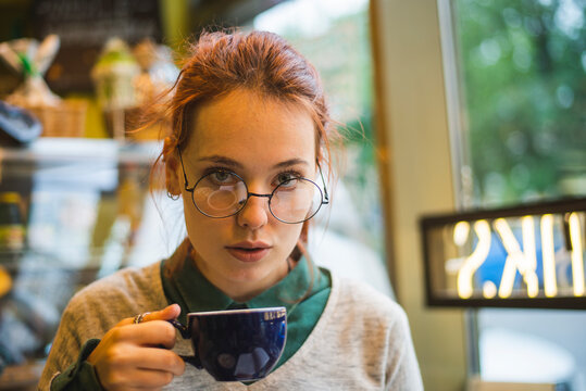 Stylish female sitting at table in cozy cafe and drinking aromatic hot beverage at weekend looking at camera