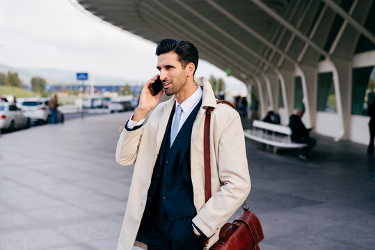 Busy man with dark hair in suit and coat speaking on phone while standing near airport terminal at daytime
