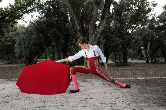 Side view of slim male bullfighter in red pants and white shirt holding cloak performing gracefully before corrida in nature