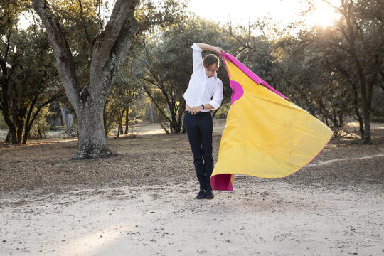 Slim male bullfighter in black pants and white shirt holding pink and yellow cloak performing gracefully before corrida in nature