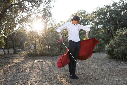 Slim focused male bullfighter in black pants and white shirt holding red cloak performing gracefully before corrida in nature