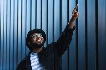 Smiling amazed hipster African American guy in hat and eyeglasses standing against wall of modern building and pointing up