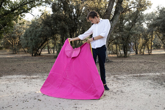 Slim male bullfighter in black pants and white shirt holding pink and yellow cloak performing gracefully before corrida in nature