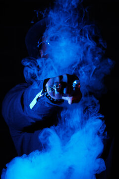 Anonymous stylish male showing fist with rings in shape of skull and covering face in cloud of smoke illuminated by blue neon light in dark studio
