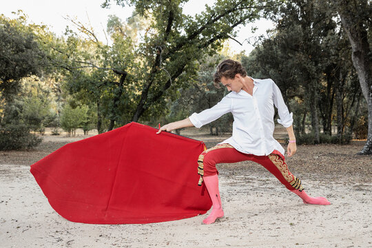 Side view of slim male bullfighter in red pants and white shirt holding cloak performing gracefully before corrida in nature